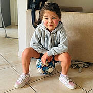 Victoire participe au concours pour gagner de l'argent avec cette photo : child, soccer_ball, indoor, hoodie, sneakers, tile_floor, smiling, young_child, person, casual_clothing, living_room, bag, toy, cute, playful, seated, sport, happy, face, curly_hair