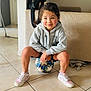 child, soccer_ball, indoor, hoodie, sneakers, tile_floor, smiling, young_child, person, casual_clothing, living_room, bag, toy, cute, playful, seated, sport, happy, face, curly_hair