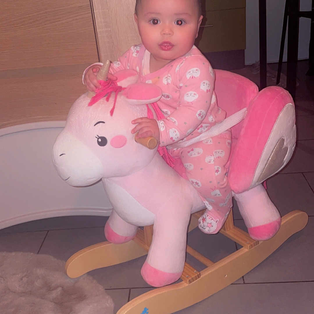 Rym a rejoint le concours — aidez-le/la à gagner de superbes lots ! baby, carpet, child, childrens_toy, cute, expression, face, floor, floor_tiles, furniture, holding, home, indoor, person, pink_pajamas, plush, rocking_unicorn, seated, toy, wooden_rocker