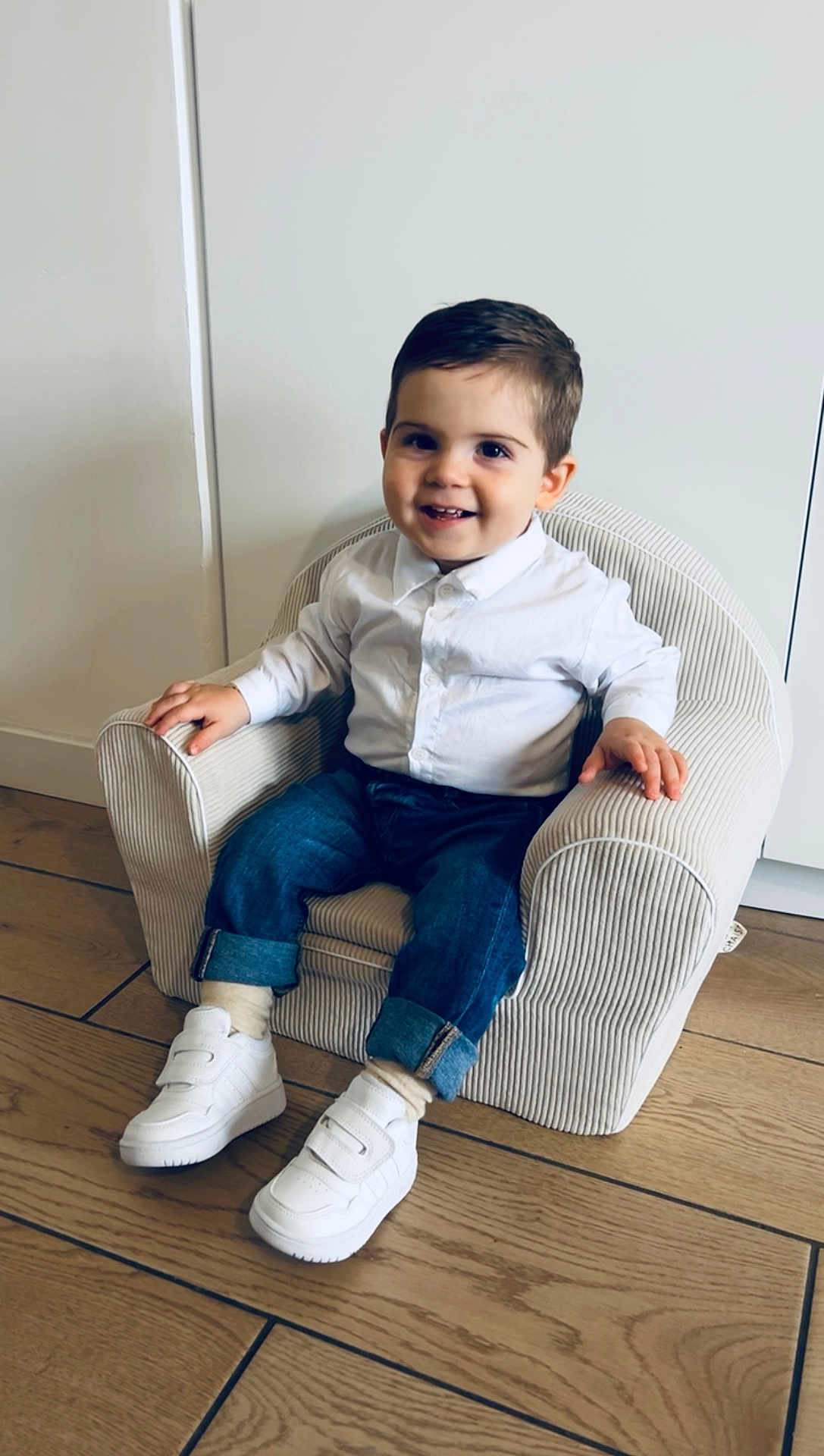 Ezio participe au concours pour gagner de l'argent avec cette photo : toddler, child, smiling, white_shirt, jeans, sneakers, armchair, striped, indoor, wooden_floor, happy, seated, casual, cute, person, young_child, furniture, portrait, small_chair, home