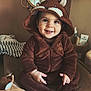baby, child, smiling, hooded_clothing, brown_onesie, fox_costume, indoor, cushion, mirror, toiletries, happy, cute, portrait, sitting, cozy, furniture, hand_jewelry, face, person, warm_lighting