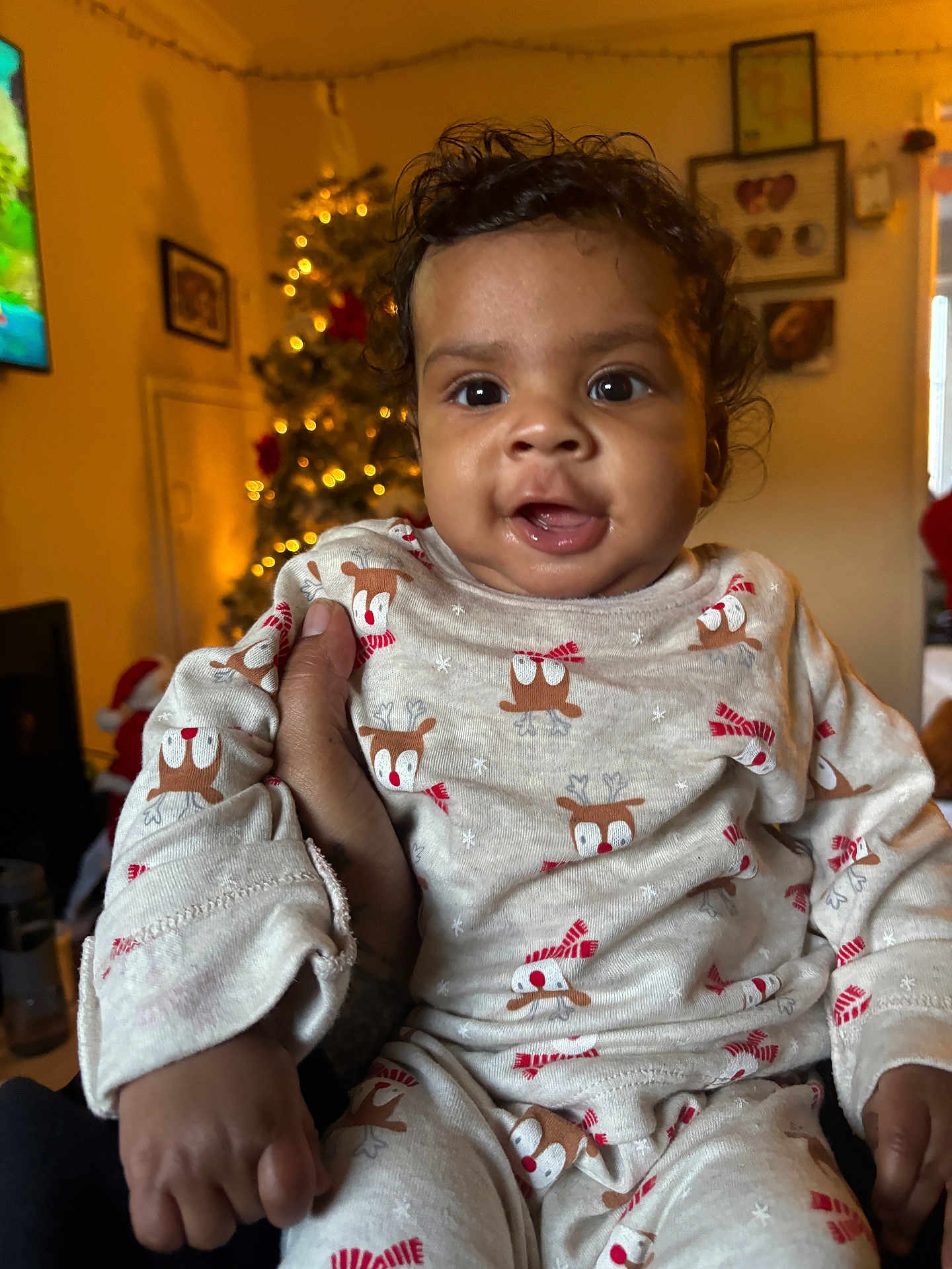 Amari is registered to the contest to win money with this photo: baby, child, infant, pajamas, reindeer_pattern, holiday, christmas_tree, indoor, hand, smiling, cute, festive, warm_lighting, person, portrait, cozy, decorations, background, holiday_season, celebration