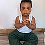 baby, barefoot, child, curly_hair, cute, floor, green_pants, hands_clasped, home, indoors, looking_at_camera, person, plain_wall, portrait, serious_expression, sitting, toddler, white_tank_top, wooden_floor, young_child