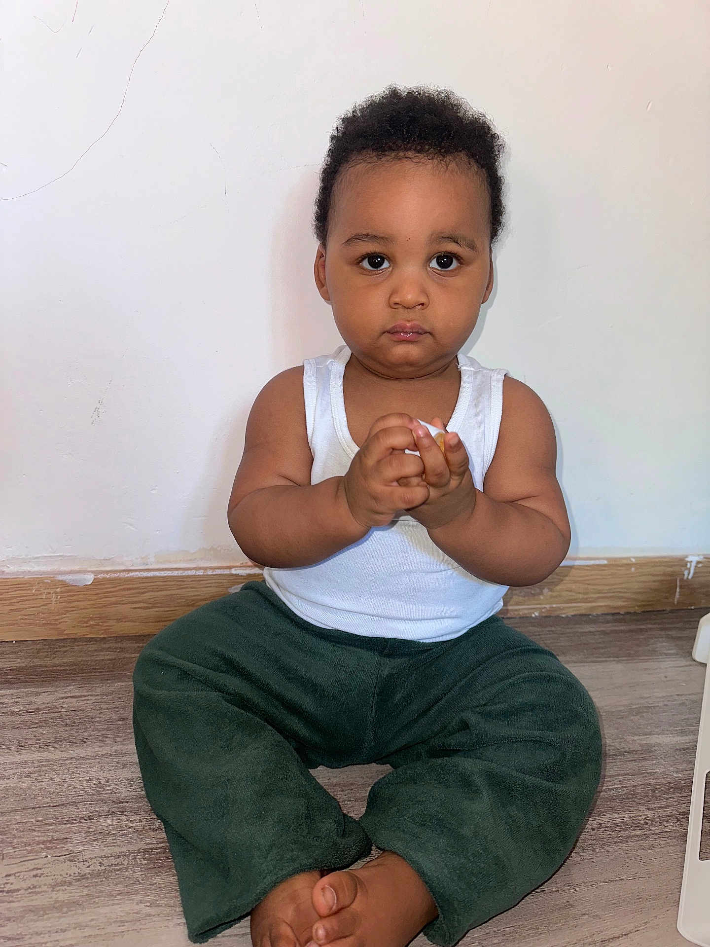Wiaam participe au concours pour gagner de l'argent avec cette photo : baby, barefoot, child, curly_hair, cute, floor, green_pants, hands_clasped, home, indoors, looking_at_camera, person, plain_wall, portrait, serious_expression, sitting, toddler, white_tank_top, wooden_floor, young_child