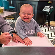 Colton joined the competition — help win amazing prizes! baby, smiling, high_chair, sweater, hand, indoor, cafe, table, person, child, happy, face, clothing, seat, adult, furniture, background, patterned_floor, holding, cute