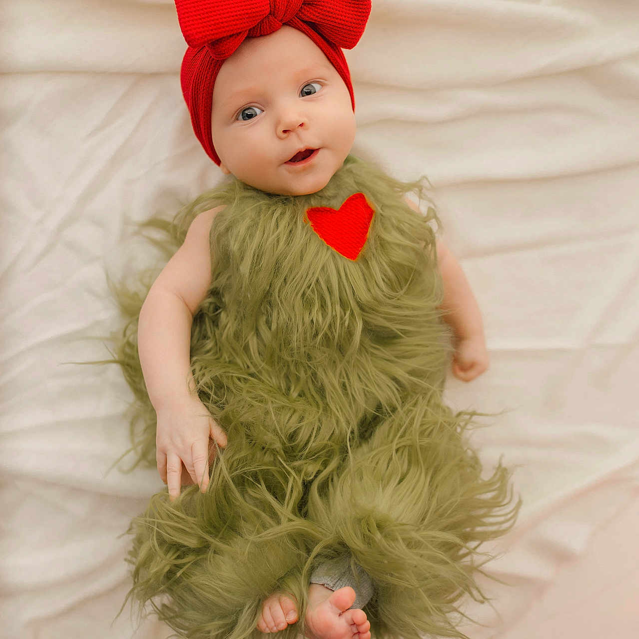 Aurora is registered to the contest to win money with this photo: adorable, baby, blanket, child, cozy, curious_expression, cute, face, green_furry_outfit, headband, heart_patch, human, indoors, infant, lying_down, portrait, red_bow, skin, small_feet, soft_texture