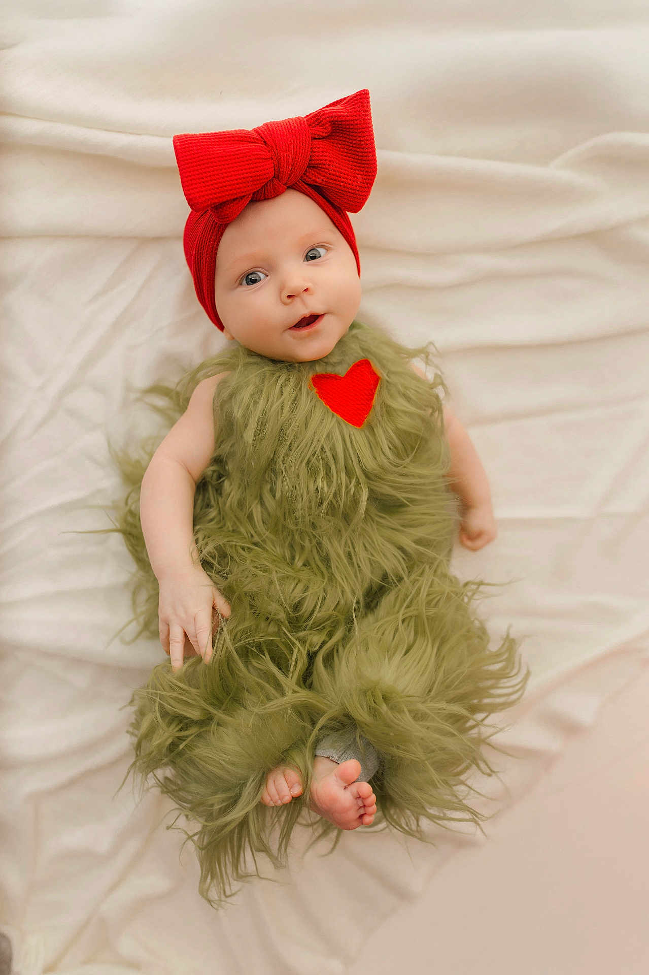 Aurora is registered to the contest to win money with this photo: baby, infant, red_bow, headband, green_furry_outfit, heart_patch, blanket, soft_texture, cute, portrait, lying_down, small_feet, curious_expression, child, human, indoors, cozy, adorable, skin, face