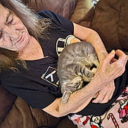 Boots is registered to the contest to win money with this photo: elderly_woman, cat, kitten, gray_tabby, couch, blanket, holiday_theme, candy_cane_pattern, resting, cozy, indoor, casual_clothing, black_shirt, wrinkled_skin, long_hair, sleeping_cat, holding, affection, relaxation, home