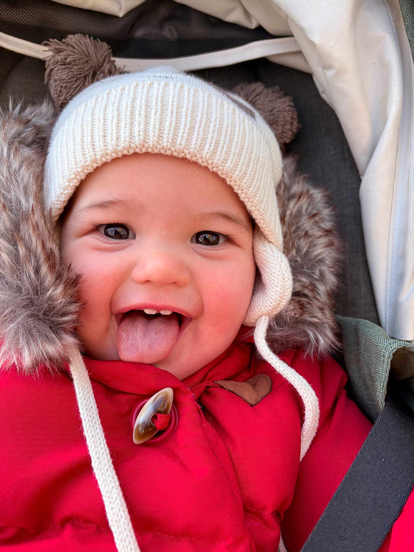 Charline a rejoint le concours — aidez-le/la à gagner de superbes lots ! baby, child, face, smile, tongue, hat, winter_clothing, red_jacket, fur_trim, cute, happy, closeup, portrait, cozy, playful, outdoor, infant, seatbelt, car_seat, childhood