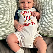 John is registered to the contest to win money with this photo: baby, infant, child, pillow, cute, smile, face, legs, socks, clothing, bed, comfortable, pattern, text, baseball, heart, hands, portrait, indoors, lying_down