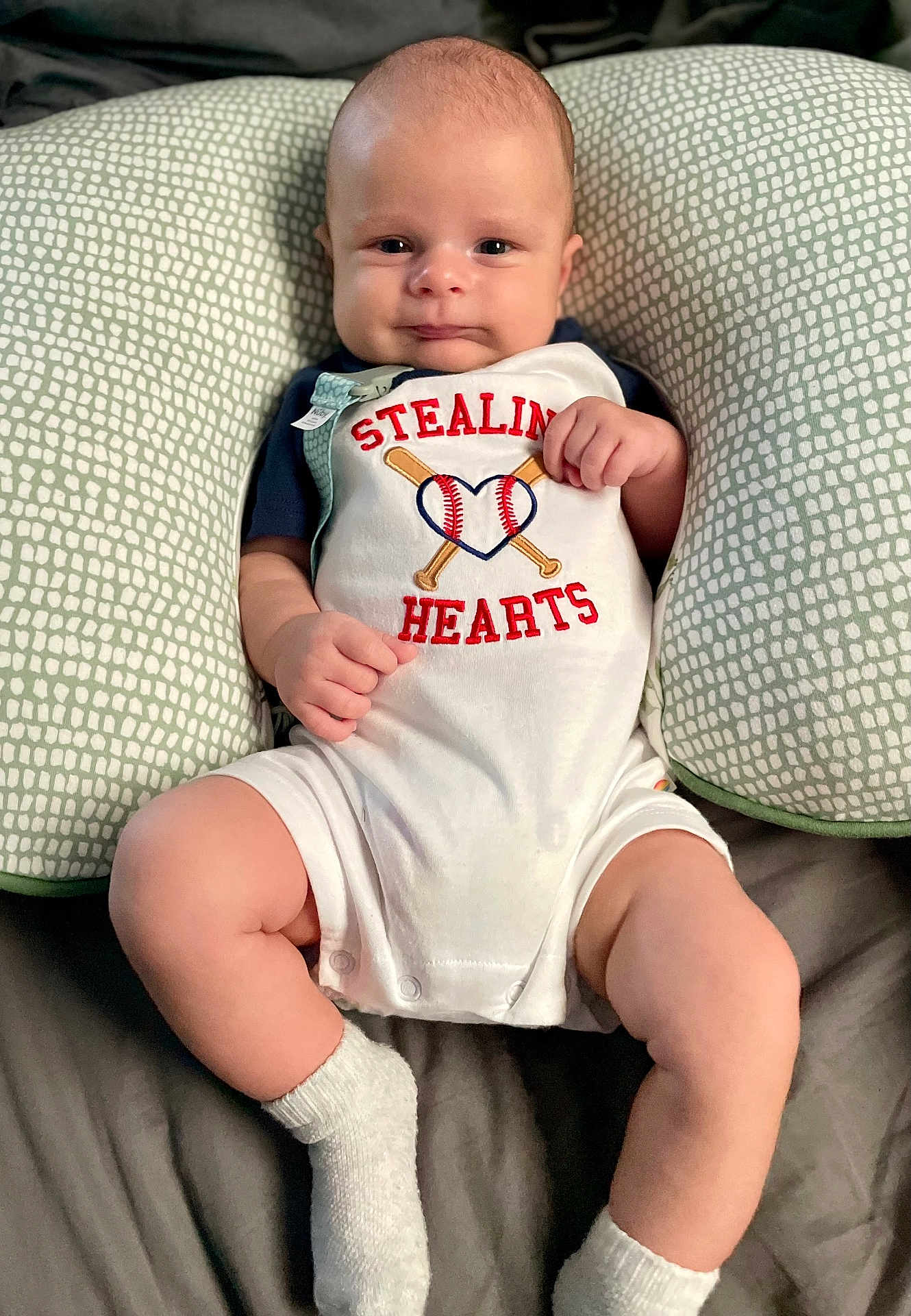 John is registered to the contest to win money with this photo: baby, infant, child, pillow, cute, smile, face, legs, socks, clothing, bed, comfortable, pattern, text, baseball, heart, hands, portrait, indoors, lying_down