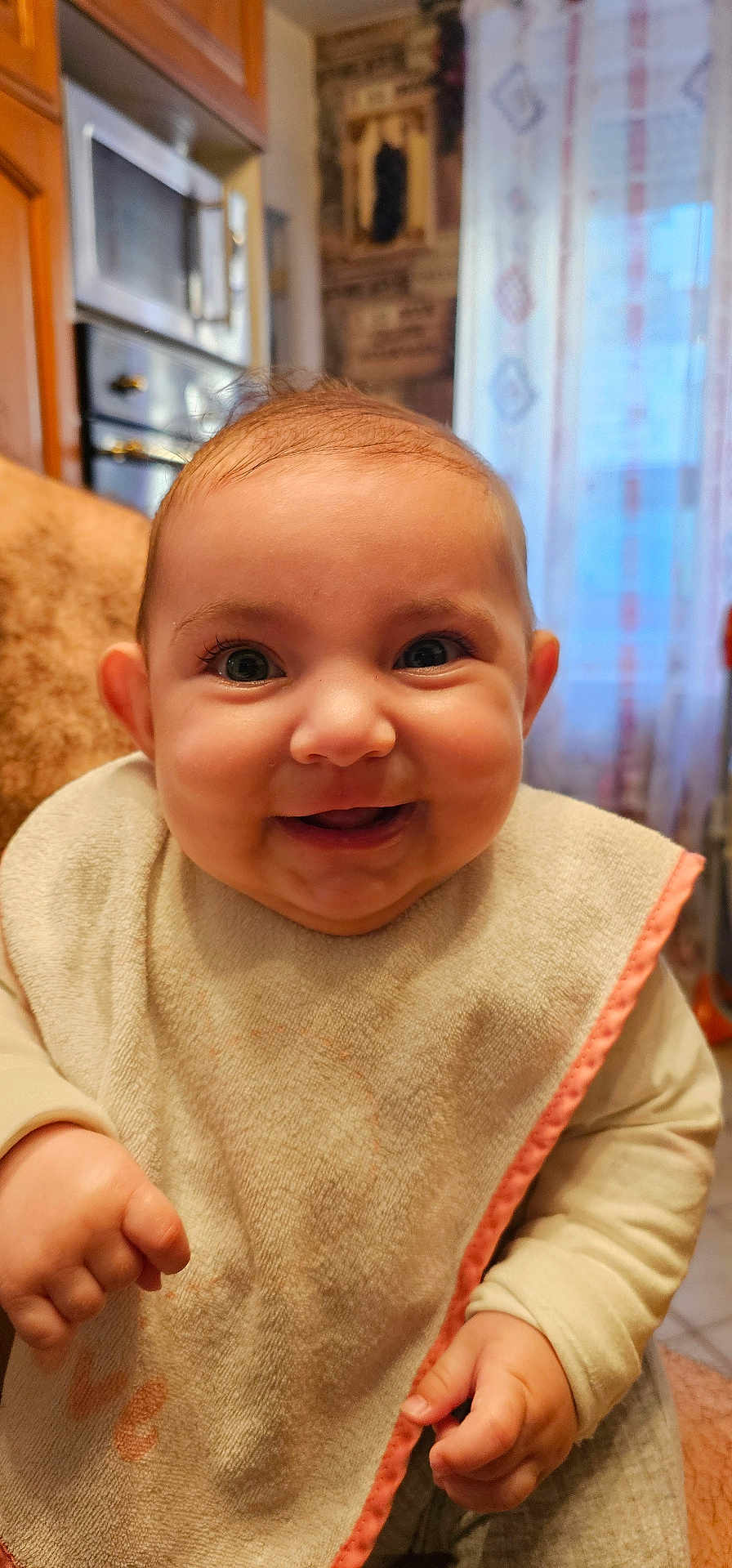 Maïlana participe au concours pour gagner de l'argent avec cette photo : baby, smiling, bib, indoor, child, cute, happy, face, hands, clothing, kitchen, window, curtain, person, infant, portrait, skin, young_child, sitting, closeup
