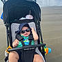 baby, stroller, beach, sunglasses, hat, plush_toy, sand, ocean, water, child, sun_hat, baby_clothing, baby_seat, toy, outdoor, sunny, relaxing, summer, person, infant