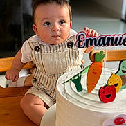 Emmanuel participe au concours pour gagner de l'argent avec cette photo : baby, child, birthday_cake, cake, table, wooden_table, overalls, striped_clothing, face, eyes, cheeks, indoor, decorations, toy_figures, vegetables, smile, celebration, person, cute, curious