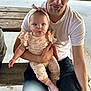 baby, man, outdoor, bench, grass, headband, white_tshirt, black_pants, playful, tongue_out, holding, infant, cute, portrait, casual, family, daylight, wooden_bench, smiling, child
