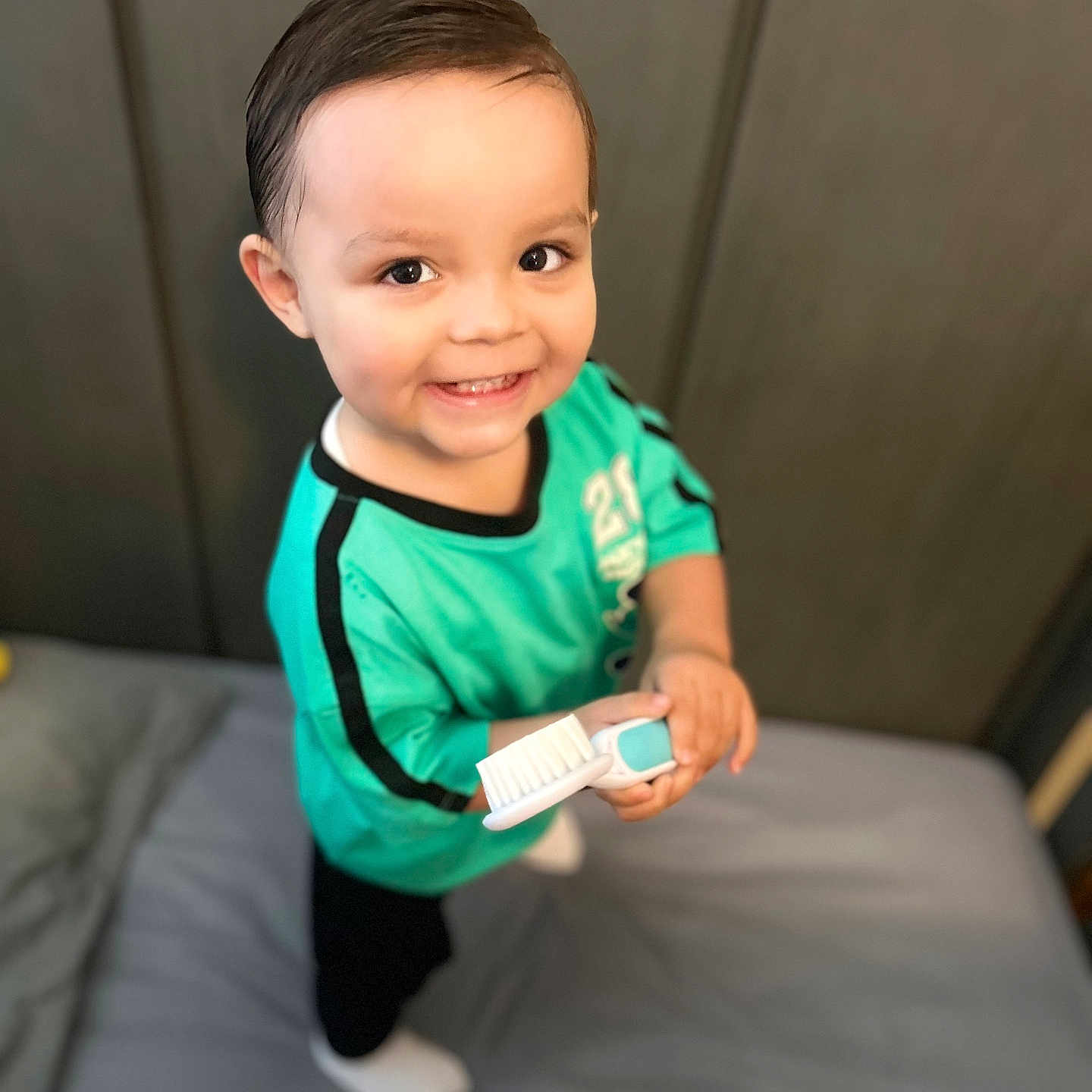 Kobe joined the competition — help win amazing prizes! toddler, child, smiling, toothbrush, bed, blanket, plush_toy, hair, green_shirt, black_pants, white_socks, indoor, person, happy, cute, portrait, room, soft_toys, furniture, playful