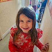 Annalya participe au concours pour gagner de l'argent avec cette photo : blurred_background, casual_clothing, child, floor, girl, hallway, happy, holding_object, home, indoor, long_hair, person, portrait, red_dress, smiling, snowflake_pattern, standing, two_people, wall, young