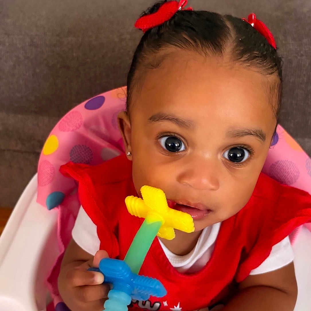 Salyn joined the competition — help win amazing prizes! baby, child, high_chair, sitting, indoor, portrait, face, wide_eyes, red_bows, hair, red_clothing, white_shirt, toy, colorful_toy, teething_toy, bracelet, couch, cushions, home, smiling