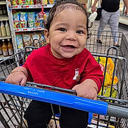 Kaleb is registered to the contest to win money with this photo: baby, child, smiling, grocery_store, shopping_cart, red_shirt, black_pants, sneakers, supermarket, shelves, food, snacks, shopping, portrait, indoor, happy, sitting, infant, adult, cart