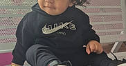 Kaleb is registered to the contest to win money with this photo: bench, black_hoodie, casual_clothing, child, curly_hair, fashion, flowers, happy, indoor, nike, photograph, pink_wall, playful, portrait, shoes, sitting, smiling, sneakers, toddler, window