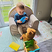 Liam participe au concours pour gagner de l'argent avec cette photo : toddler, child, armchair, toy, blue_toy, teddy_bear, wooden_toy_box, play_mat, indoor, window, patio_chair, curious, sitting, floor, foot, clothing, person, baby, home, daylight
