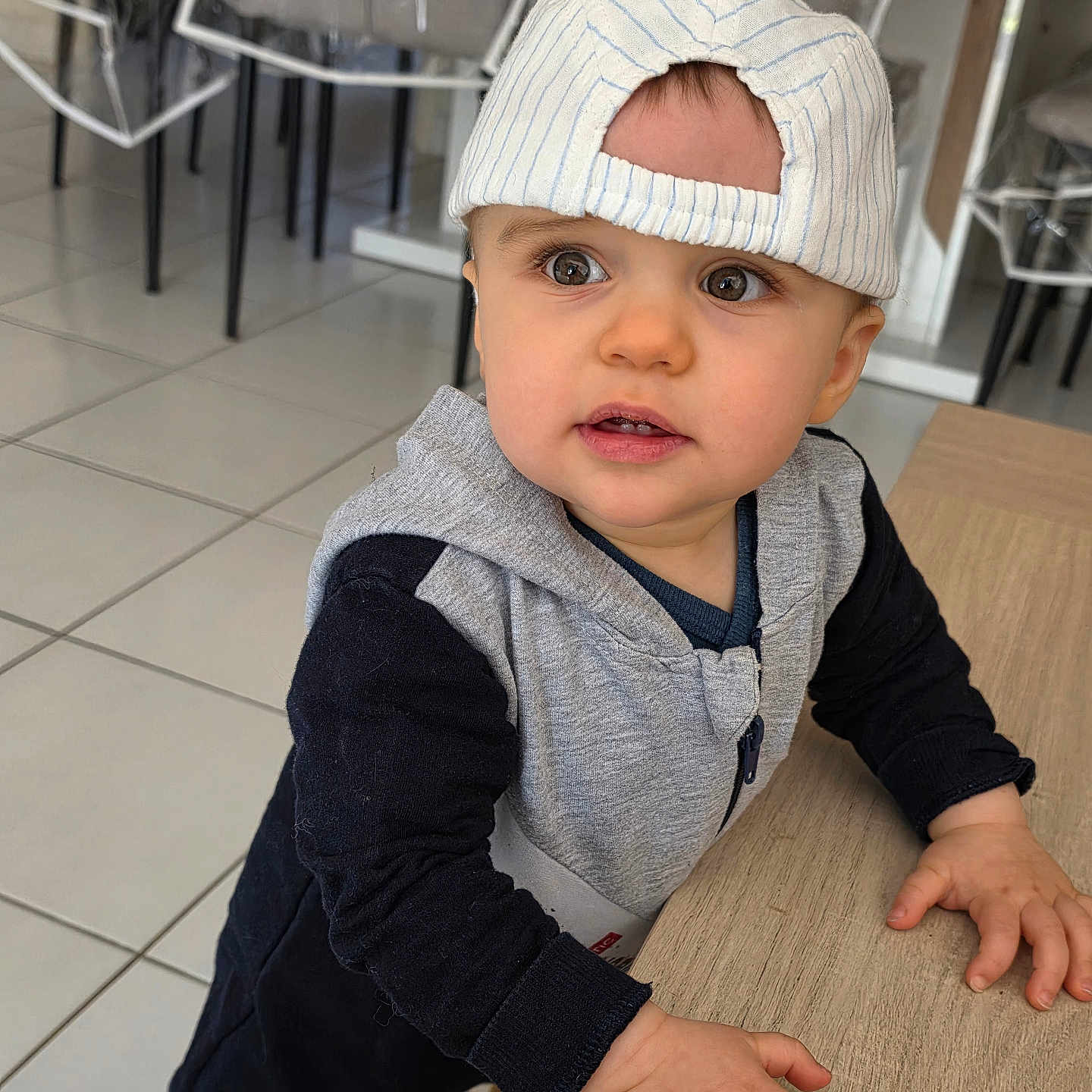 Liam a rejoint le concours — aidez-le/la à gagner de superbes lots ! toddler, child, baby, cap, striped_cap, table, hands, indoor, floor, tiles, curious, eyes, face, clothing, hoodie, long_sleeve, person, leaning, furniture, chair