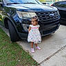 child, girl, dress, shoes, car, ford, explorer, driveway, sidewalk, grass, speed_limit_sign, outdoor, vehicle, concrete, parking, curb, tree, sky, daylight, casual