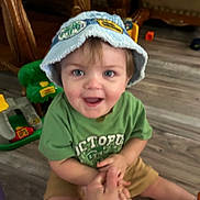 Jameson is registered to the contest to win money with this photo: toddler, child, hat, blue_hat, green_shirt, smiling, sitting, hand_holding, toy, wooden_floor, indoor, furniture, playtime, happy, person, face, shorts, casual_clothing, floor, playroom