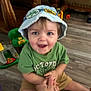 toddler, child, hat, blue_hat, green_shirt, smiling, sitting, hand_holding, toy, wooden_floor, indoor, furniture, playtime, happy, person, face, shorts, casual_clothing, floor, playroom