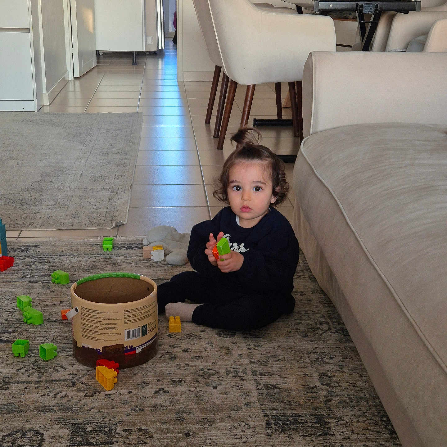 Isra a rejoint le concours — aidez-le/la à gagner de superbes lots ! building_blocks, casual_clothing, chairs, child, container, curly_hair, daylight, floor, glass, home, indoor, living_room, person, playing, rug, sofa, table, toddler, topknot, toys