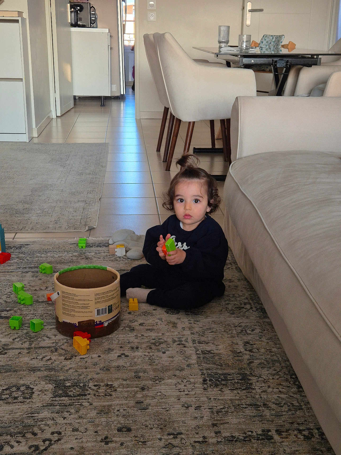 Isra a rejoint le concours — aidez-le/la à gagner de superbes lots ! building_blocks, casual_clothing, chairs, child, container, curly_hair, daylight, floor, glass, home, indoor, living_room, person, playing, rug, sofa, table, toddler, topknot, toys