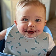 Cooper is registered to the contest to win money with this photo: baby, child, high_chair, bib, blueberries, food_stain, face, smiling, indoor, kitchen, young_child, cute, messy, feeding, person, close_up, table, seat, whale_pattern, happy