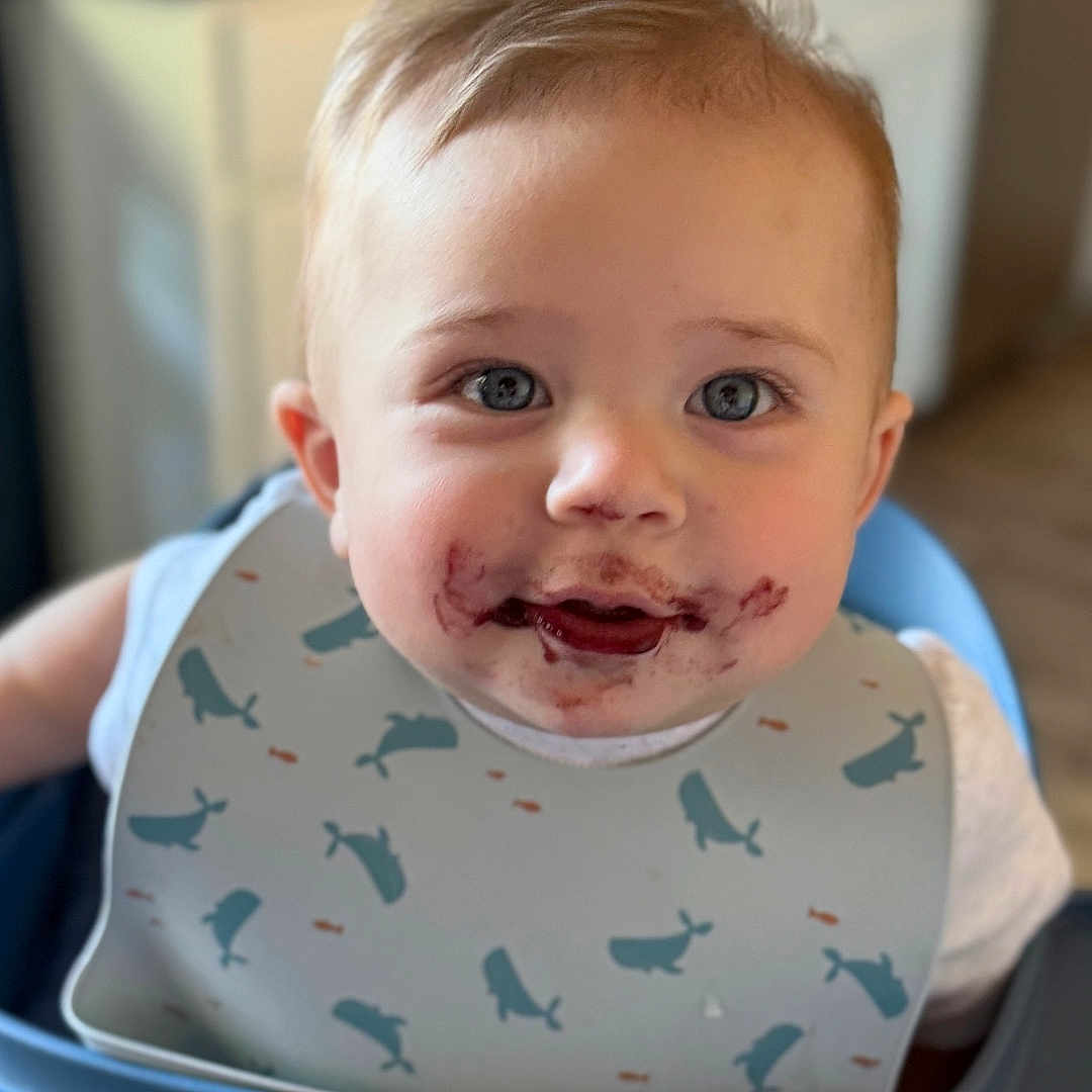 Cooper is registered to the contest to win money with this photo: baby, child, high_chair, bib, blueberries, food_stain, face, smiling, indoor, kitchen, young_child, cute, messy, feeding, person, close_up, table, seat, whale_pattern, happy