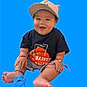 baby, child, cap, smiling, black_shirt, basketball_graphic, blue_toy, sitting, happy, cute, footwear_none, bracelets, shorts, indoor, portrait, playful, infant, bright_background, isolated, studio_style