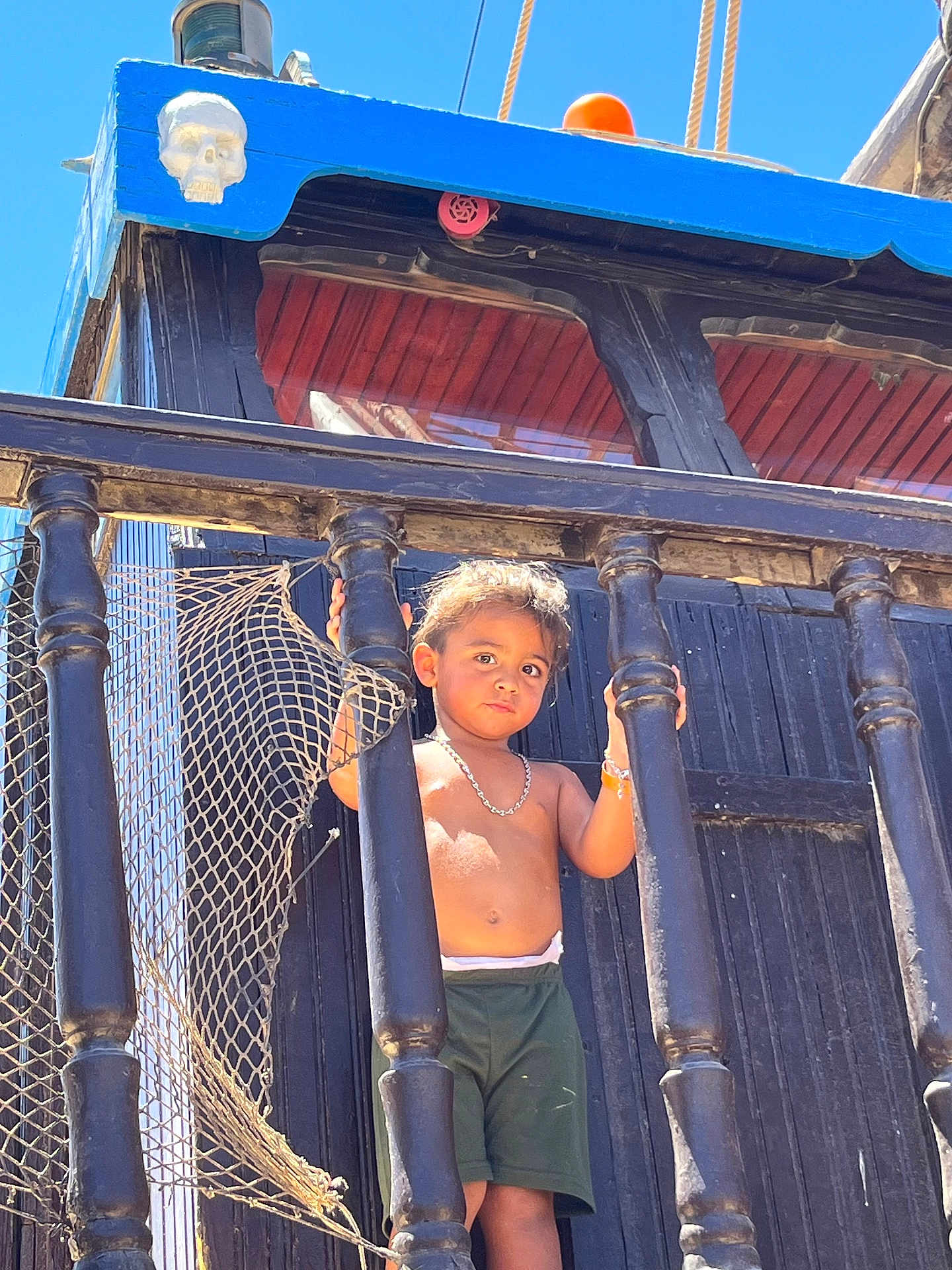 Naim Fridhi a rejoint le concours — aidez-le/la à gagner de superbes lots ! child, toddler, boy, shirtless, shorts, bracelet, necklace, wooden_railings, netting, pirate_ship, skull_decoration, blue_sky, outdoor, sunlight, daytime, portrait, serious_expression, standing, playground, adventure