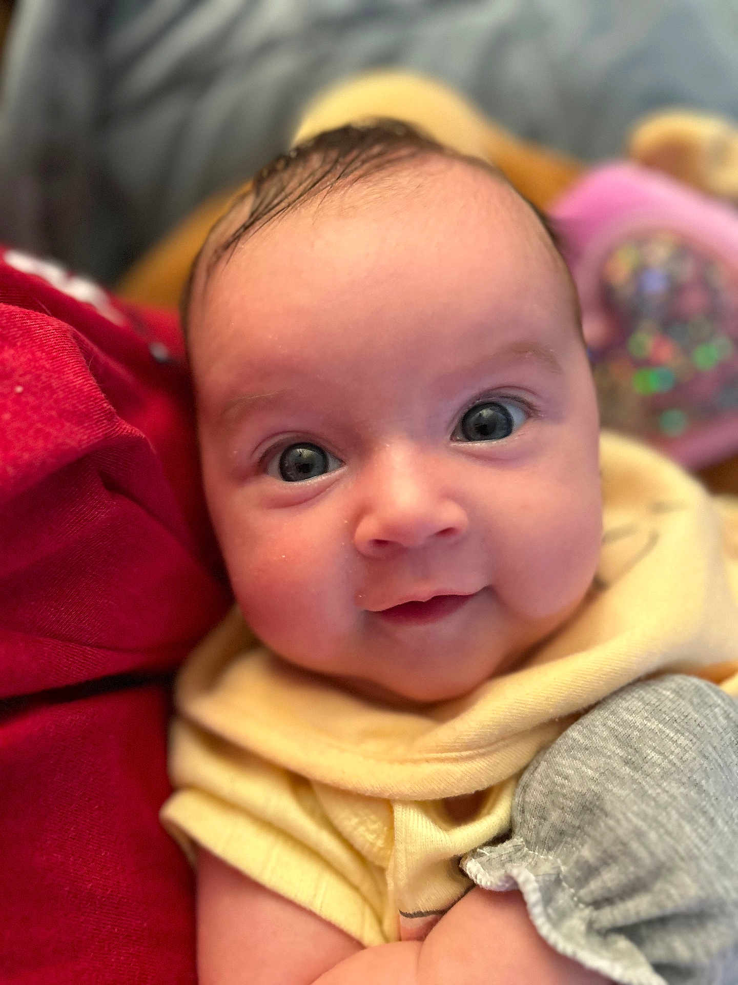 Evie is registered to the contest to win money with this photo: baby, infant, face, eyes, smile, yellow_clothing, gray_clothing, red_fabric, close_up, cute, skin, head, person, child, portrait, newborn, soft_lighting, indoors, expression, warm