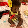 toddler, child, bed, yellow_flower, curly_hair, red_card, clothing, indoors, person, skin, sitting, barefoot, yellow_top, bedroom, blanket, soft_lighting, cute, focus, young_child, hair_accessory