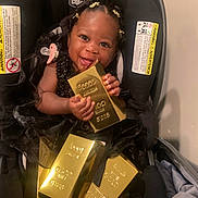 Azmariyah is registered to the contest to win money with this photo: baby, child, car_seat, black_dress, gold_bars, smile, happy, indoor, pacifier, hair_clips, person, cute, infant, seat, holding, shiny, luxury, portrait, young, adorable
