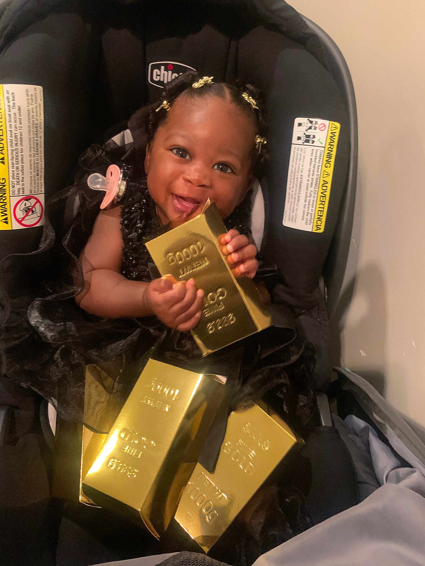 Azmariyah is registered to the contest to win money with this photo: baby, child, car_seat, black_dress, gold_bars, smile, happy, indoor, pacifier, hair_clips, person, cute, infant, seat, holding, shiny, luxury, portrait, young, adorable