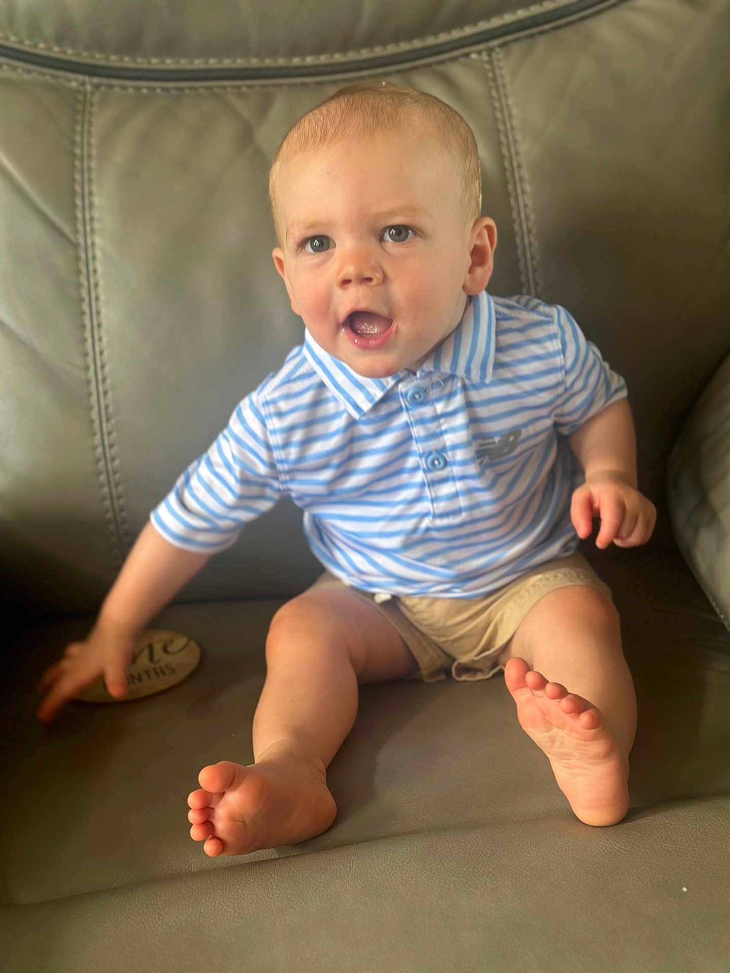 Darren is registered to the contest to win money with this photo: baby, child, polo_shirt, shorts, sitting, couch, leather, foot, hand, face, happy, smiling, blue, striped, skin, indoors, person, young_child, expression, casual_clothing