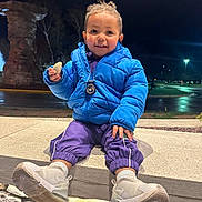 Shai is registered to the contest to win money with this photo: blue_jacket, casual_clothing, child, curly_hair, cute, happy, holding_rock, night, outdoor, person, portrait, purple_pants, sitting, smiling, stone_wall, street_lights, toddler, urban, warm_clothing, white_sneakers