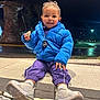 blue_jacket, casual_clothing, child, curly_hair, cute, happy, holding_rock, night, outdoor, person, portrait, purple_pants, sitting, smiling, stone_wall, street_lights, toddler, urban, warm_clothing, white_sneakers