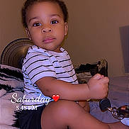 Jah'Seil joined the competition — help win amazing prizes! bed, black_shorts, casual_clothing, child, curious, cute, expression, fan, furniture, holding_object, home, indoor, person, portrait, sitting, striped_shirt, timestamp, toddler, wall, young_child