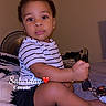 bed, black_shorts, casual_clothing, child, curious, cute, expression, fan, furniture, holding_object, home, indoor, person, portrait, sitting, striped_shirt, timestamp, toddler, wall, young_child