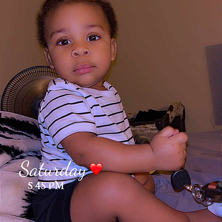 Jah'Seil joined the competition — help win amazing prizes! bed, black_shorts, casual_clothing, child, curious, cute, expression, fan, furniture, holding_object, home, indoor, person, portrait, sitting, striped_shirt, timestamp, toddler, wall, young_child