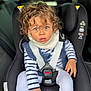 child, toddler, curly_hair, car_seat, safety_harness, striped_shirt, white_pants, scarf, seatbelt, car_interior, young_child, portrait, expressive_eyes, casual_clothing, indoor, seat, baby, person, cute, curious