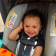 Dariyan joined the competition — help win amazing prizes! baby, child, car_seat, smile, curly_hair, toy, seatbelt, infant, happy, sitting, closeup, indoor, person, cute, playful, warning_label, black_clothing, bright_colors, face, portrait