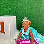baby, balloon, blue_dress, celebration, child, cute, dress, fluffy_rug, green_background, happy, holding, indoor, infant, number_one, party, pink_balloon, portrait, smiling, tiara, white_box