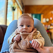 Maybel joined the competition — help win amazing prizes! baby, child, infant, person, overalls, hand, finger, lap, clothing, indoor, window, blurred_background, face, blue_eyes, cute, cozy, casual, portrait, home, seated
