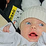 baby, infant, face, blue_eyes, hat, pom_poms, knitwear, white_clothing, hand, car_seat, airbag_warning, expression, surprised, curious, closeup, portrait, child, cute, cozy, warm
